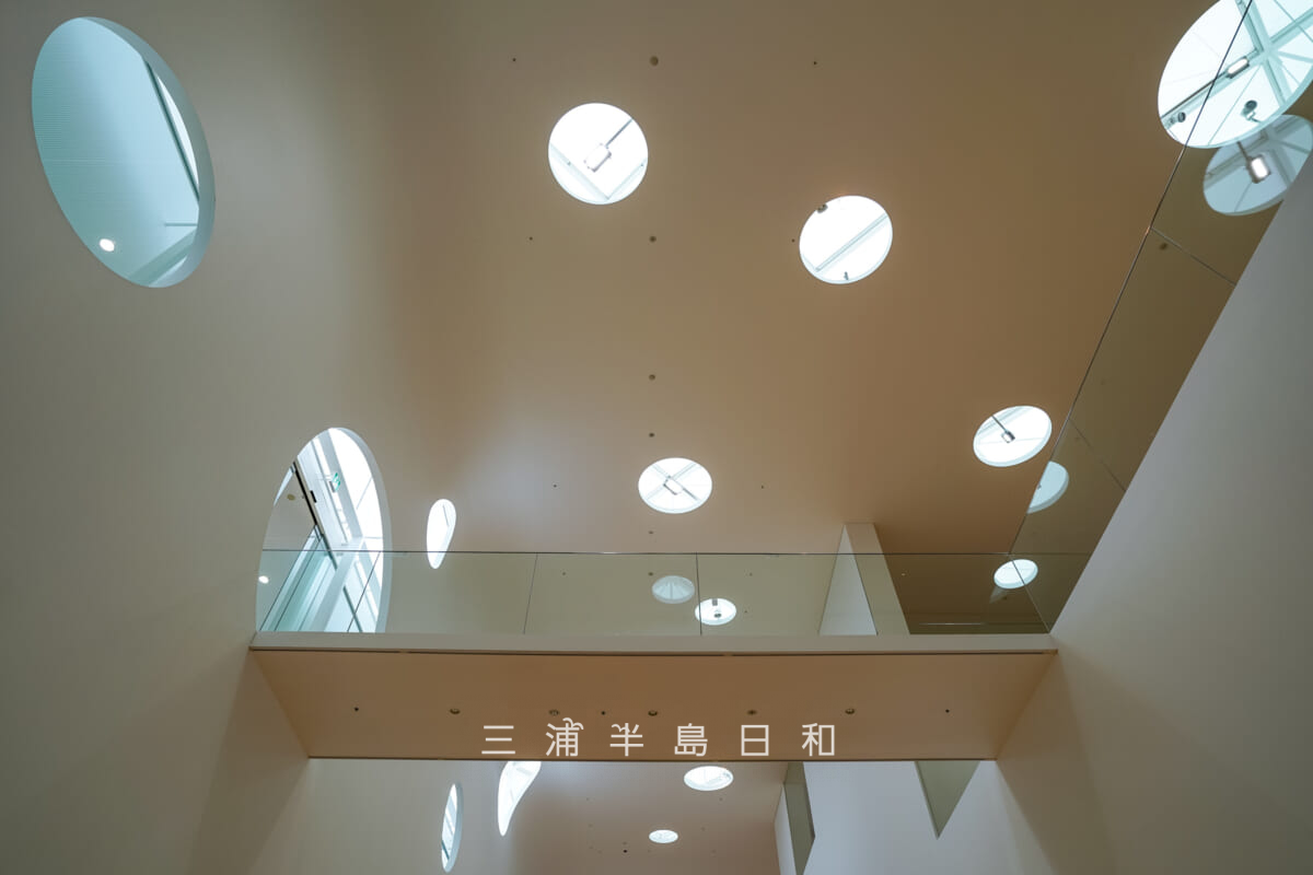 横須賀美術館・地階回廊よりランダムな丸窓が特徴的なエントランスホール前の吹き抜けを見上げる(撮影日:2024.07.01)